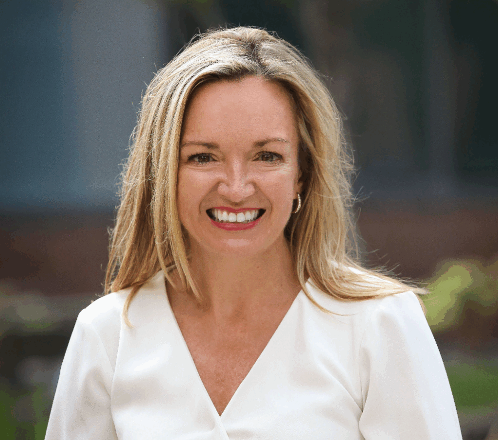 A woman with long blonde hair, wearing a white top, smiles warmly at the camera. The background is softly blurred with hints of greenery and a building.